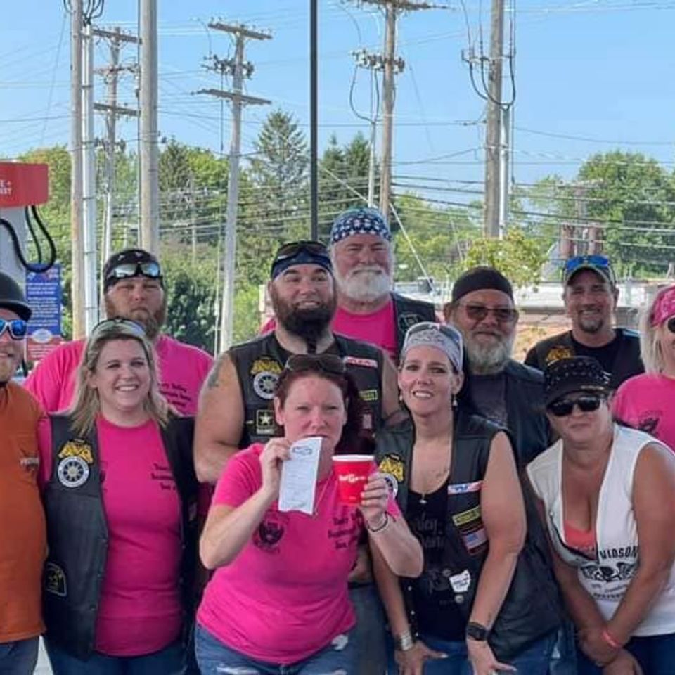 Group of motorcycle riders and supporters wearing pink event shirts posing together outdoors.