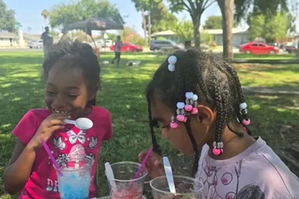 One young girl is eating a blue slushie, and another girl is eating a red slushie at the park, and they are sitting at a table together