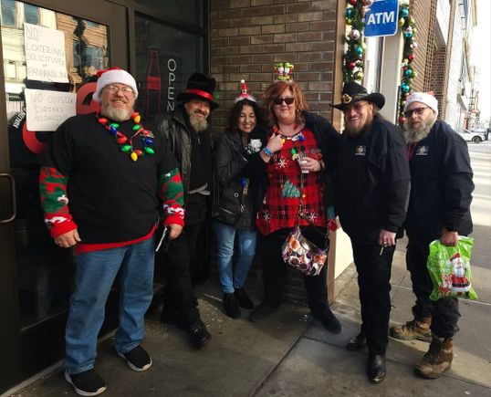 Group of event attendees wearing Christmas-themed outfits and accessories while posing on a downtown sidewalk.