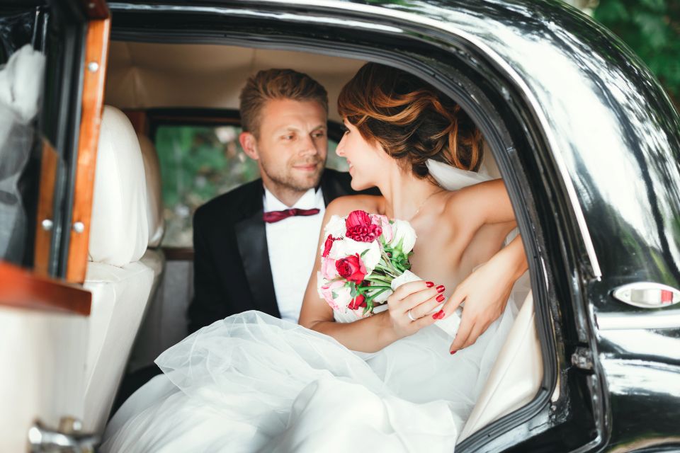 Bride and groom sitting in wedding car, looking at each other, smiling and holding bouquet