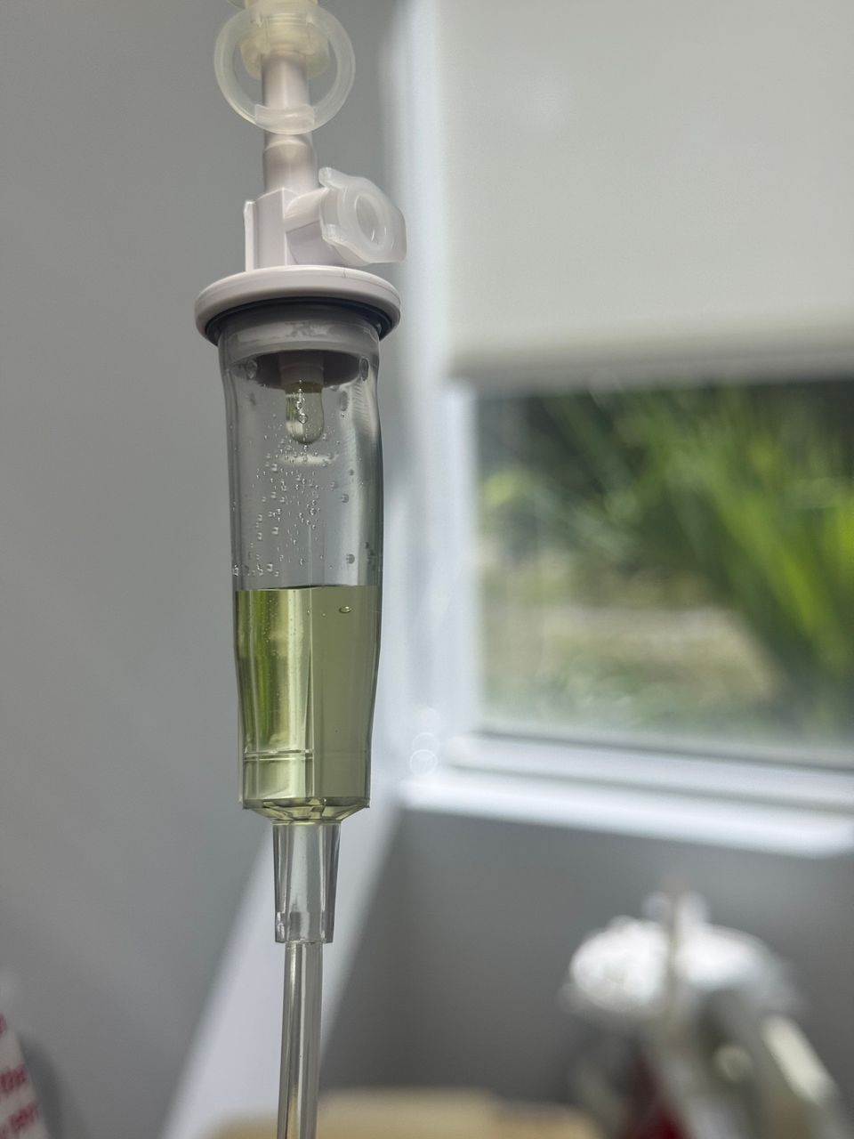 Close up of an IV drip chamber with yellow vitamin fluid beside a bright window