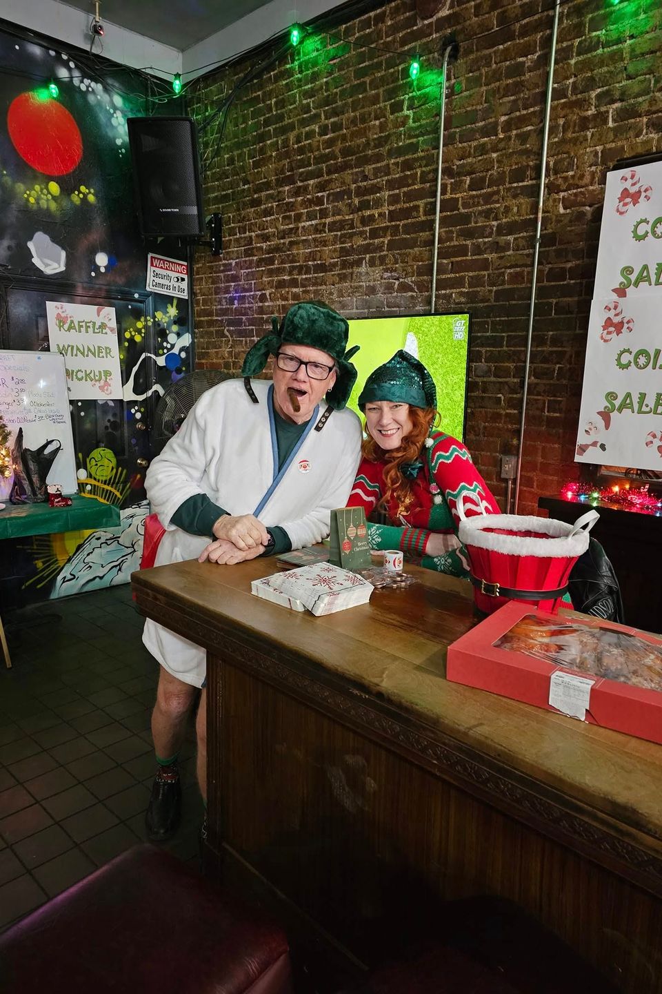 Two people in festive holiday outfits standing behind a table at a Christmas-themed fundraiser.