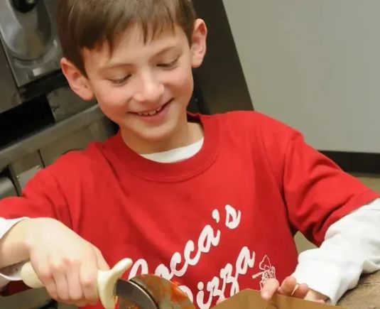 Child wearing a Cocca’s Pizza shirt preparing a personal pizza.
