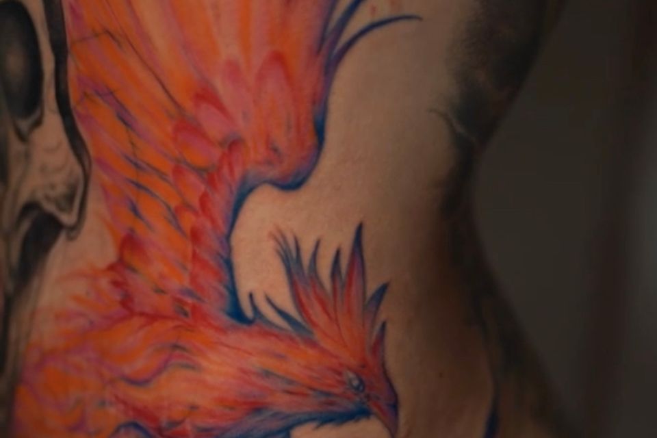 Image of a large colorful phoenix tattoo on a womans left side just under the breast