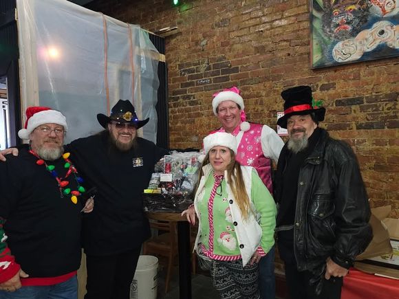 Group of attendees in holiday attire posing indoors beside raffle baskets during a Christmas-themed gathering.