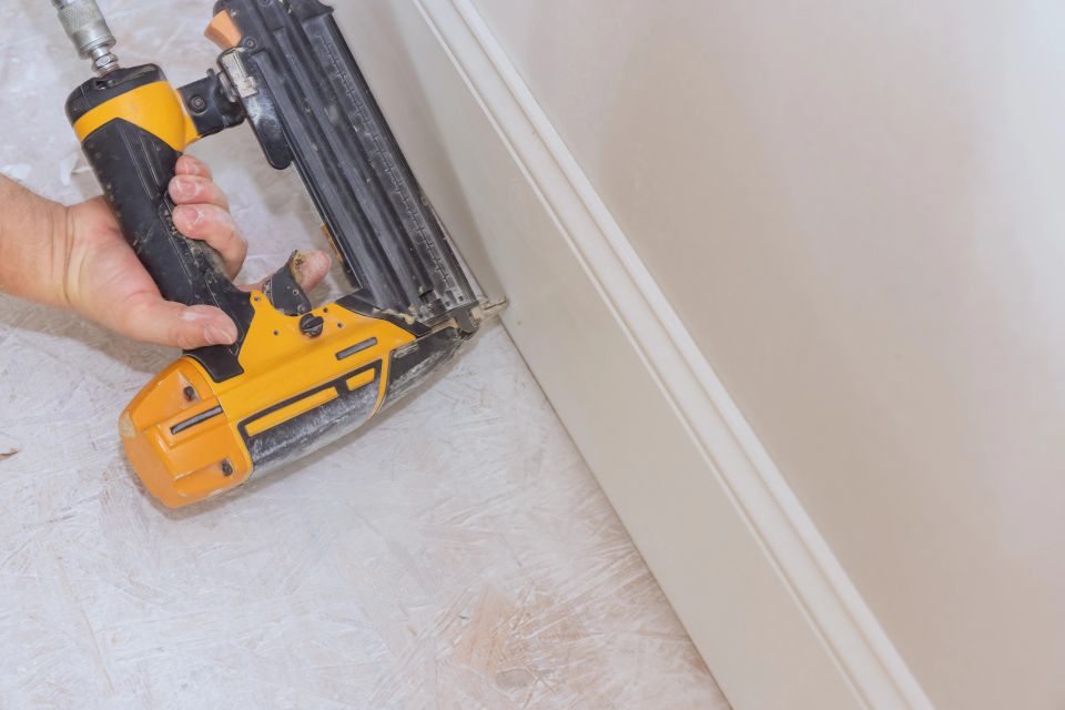 Installer using a brad nail gun to fasten white baseboard trim to the wall