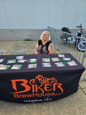 Woman seated behind a promotional table with merchandise at an outdoor event.