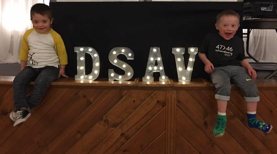 Two children with Down syndrome sitting beside illuminated DSAV letters during a Down Syndrome Association of the Valley event.
