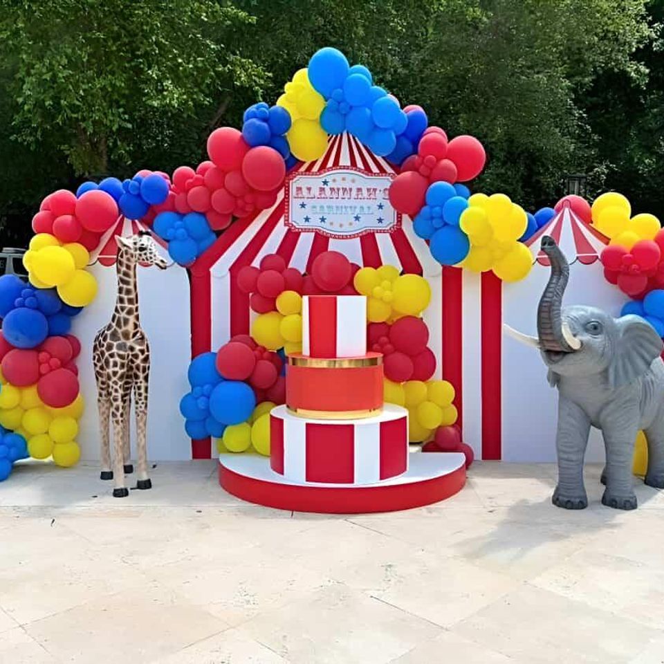Circus-themed event backdrop with red and white tent design, colorful balloon garlands, giraffe and elephant props by Aldys Events.