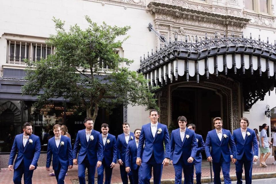 Groom and groomsmen wearing coordinated royal blue suits from New Orleans Tuxedo Inc., walking together in front of a historic New Orleans building.