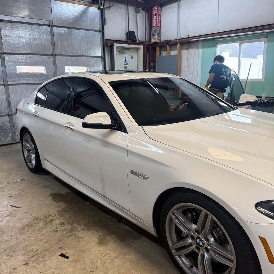 White BMW sedan with dark tinted windows as an installer works in the background of the tint shop