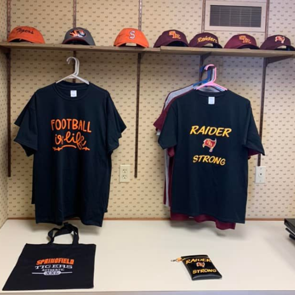 Display of custom school spirit shirts, hats, tote bag, and accessories featuring football and Raider-themed designs.