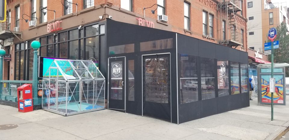 Long black framed seasonal vestibule running along a storefront and wrapping the corner, with a clear paneled enclosure beside it on the sidewalk