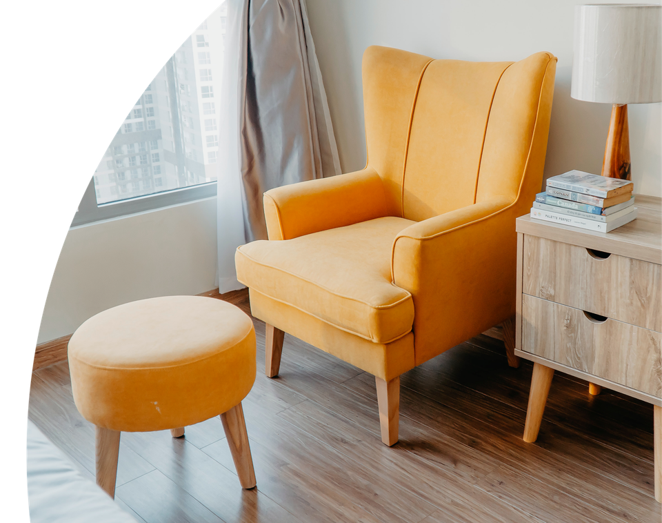 Mustard yellow accent chair with matching ottoman beside a wooden nightstand and lamp in a bright, modern living room.