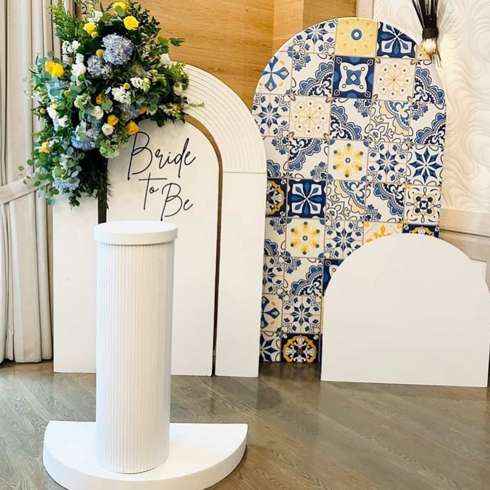Elegant bridal shower setup with ‘Bride to Be’ arch, floral arrangement, patterned tile backdrop, and white display pedestal