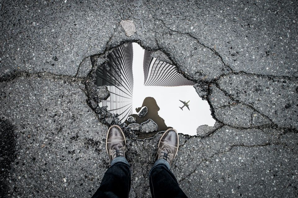 A reflection of sky scrapers and a plane flying over in a pot hole with water. Photographers feet on the edge. 
