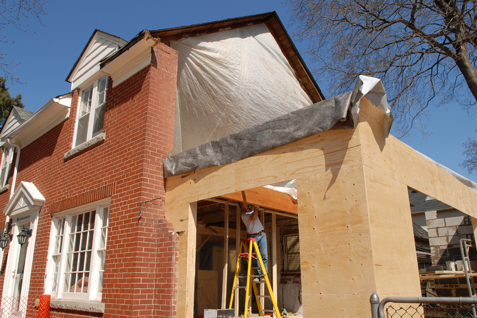 Home addition under construction featuring new framing, protective coverings, and ongoing exterior building work attached to an existing brick home.