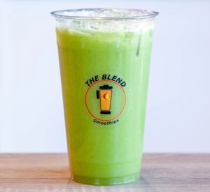 A vibrant yellow-green juice in a clear cup labeled "The Blend Smoothies," with a whole orange in front, representing a mix of apple, cucumber, ginger, and lemon.