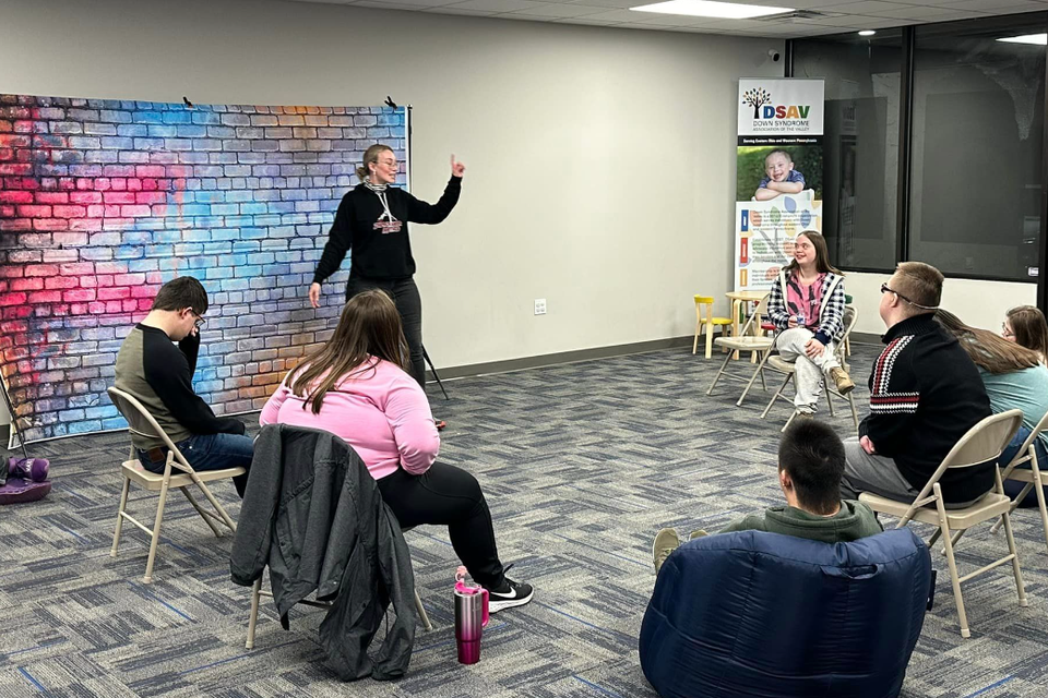 Instructor leading an improv class while participants with Down syndrome engage in theater activities in a classroom.