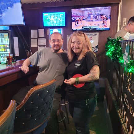 Two people posing inside a venue during a social event, with televisions and festive decorations in the background.