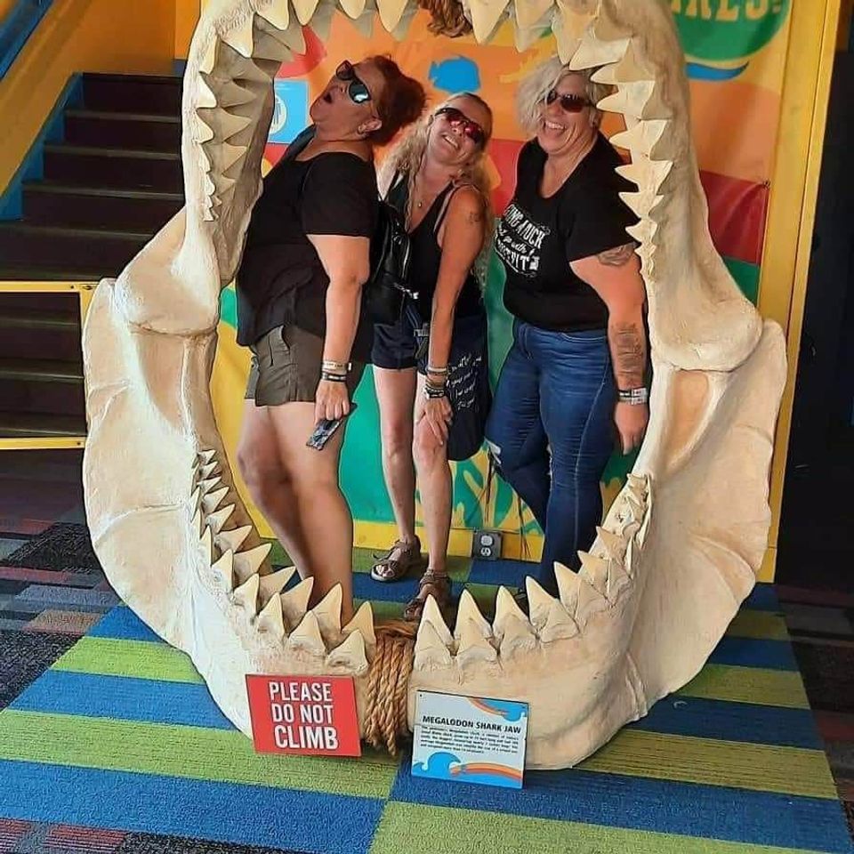 Three women smiling and posing inside a large shark jaw exhibit at an attraction.