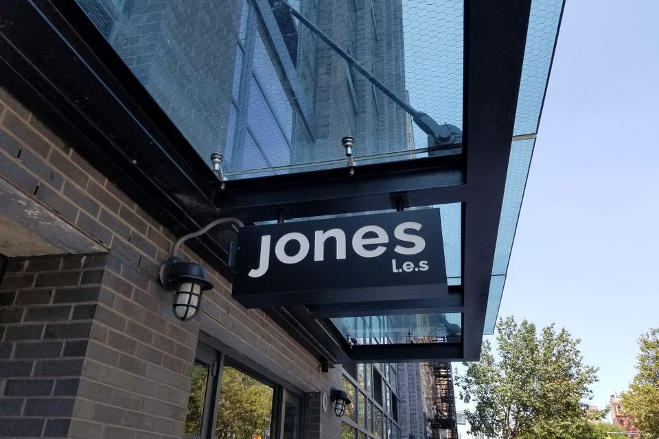 Black blade sign reading Jones L.E.S. hanging beneath a glass entrance canopy beside a wall light on the Lower East Side