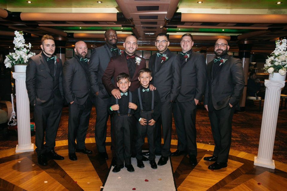 Groom, groomsmen, and two young ring bearers wearing matching dark suits posing indoors at a wedding.
