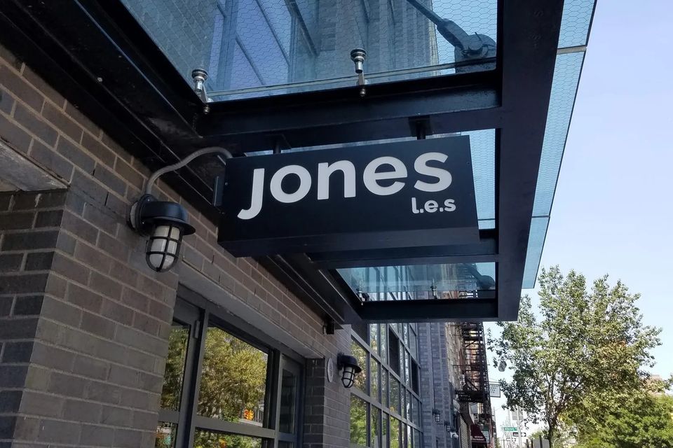 Black blade sign reading Jones L.E.S. hanging beneath a glass entrance canopy beside a wall light on the Lower East Side