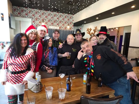 Group of attendees wearing Christmas-themed clothing and accessories gathered around a table at a holiday party indoors.