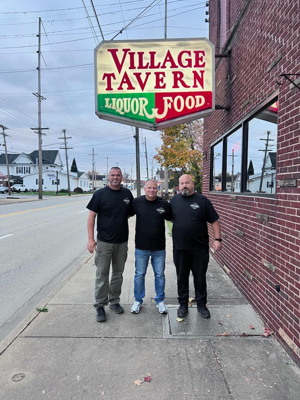 Three owners standing outside Village Tavern under the hanging sign on a city street