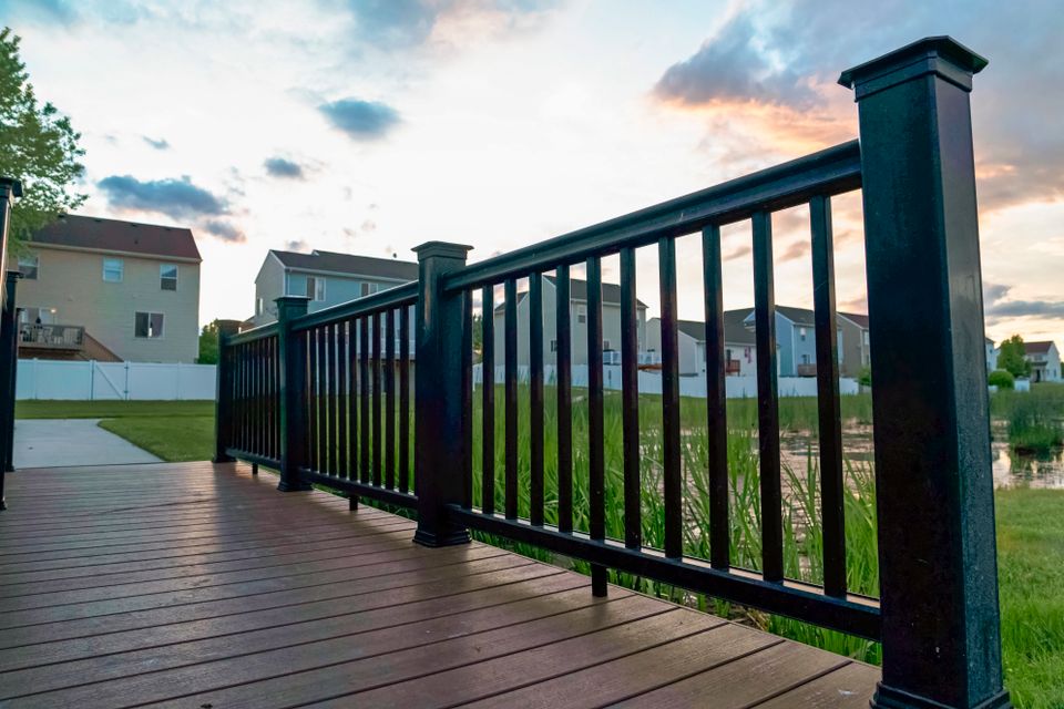 Bridge over a pond with multi storey homes and cloudy sky in the background. The structure has a brown wood deck and black guardrail