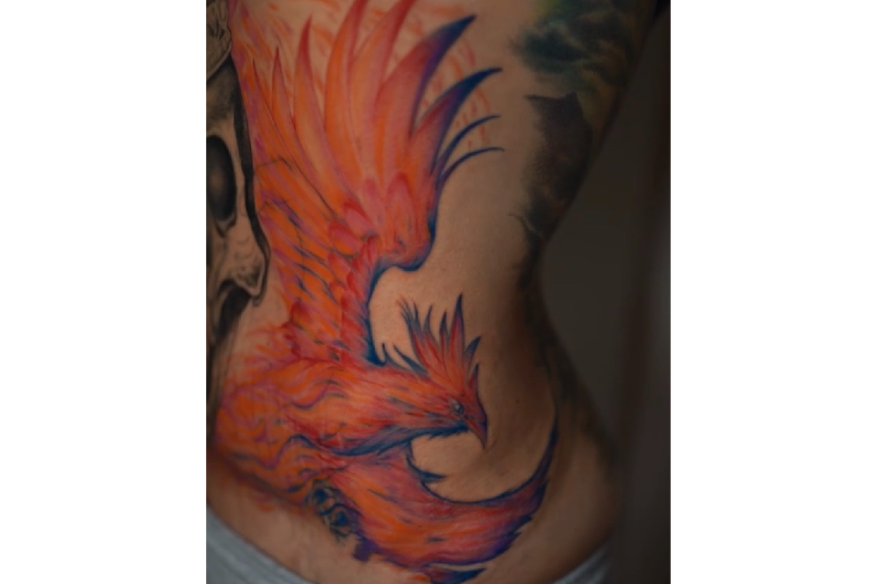Image of a phoenix in bright orange red and blue making it look like the bird is on fire. This tattoo covers a wormans left front side from just under her breast dow to her hip