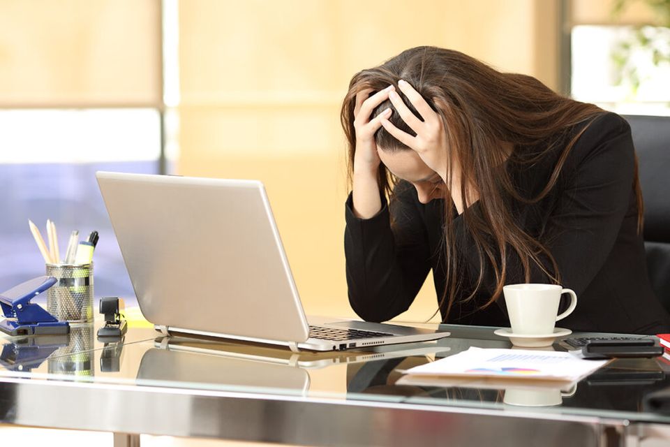 A person sitting at a desk in an office, holding their head in both hands in a gesture of stress or overwhelm, surrounded by a laptop, a cup of coffee, pens, a stapler, and a document with a colorful chart.
