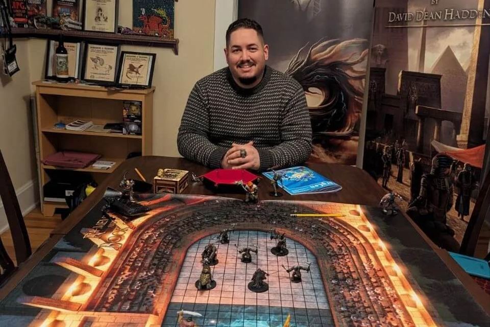 Dungeon master sitting at a game table with a detailed fantasy RPG map and miniatures, surrounded by RPG books and Orthané Productions banners