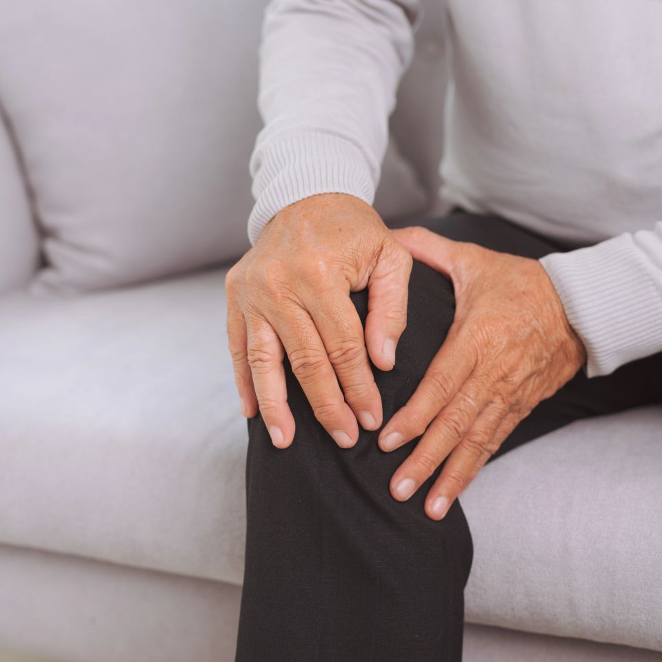 Senior man sitting on sofa in living room at home and touching his knee by the pain