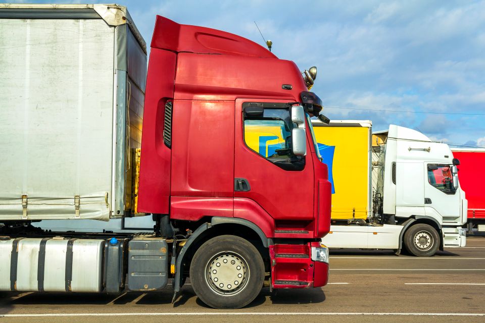 Colorful modern big semi-trucks and trailers of different makes and models stand in row on flat parking lot of truck stop in sunshine