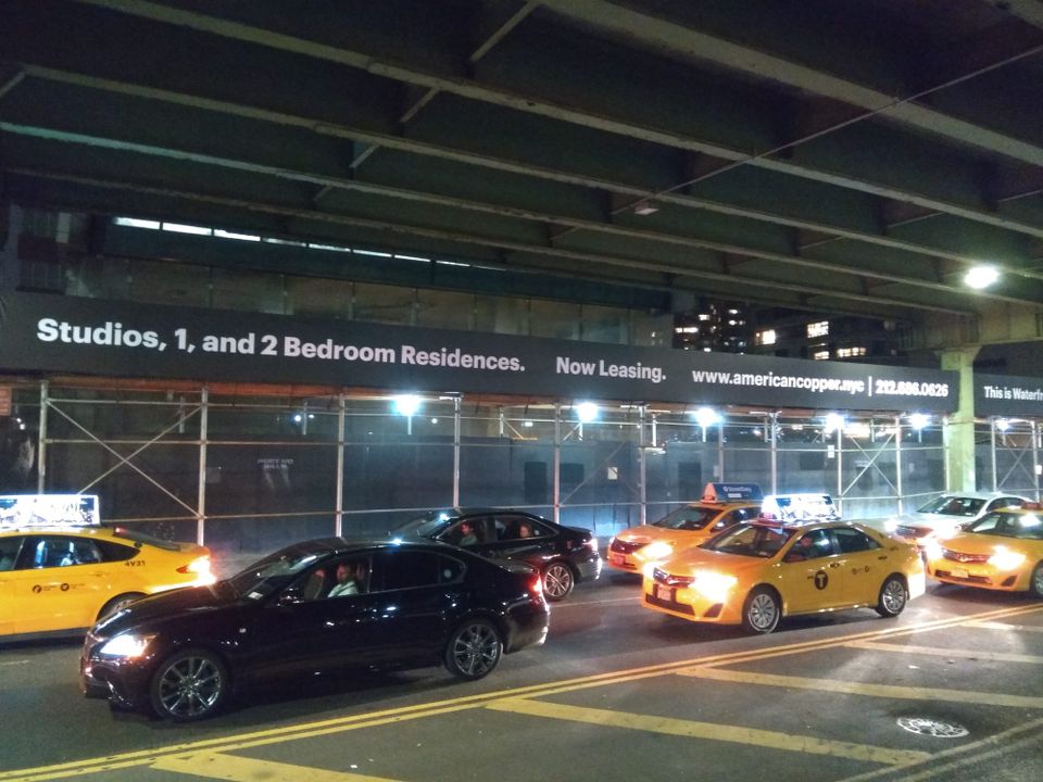 Long printed leasing banner for American Copper running along a lit scaffold beneath an elevated roadway at night, with taxis passing in front