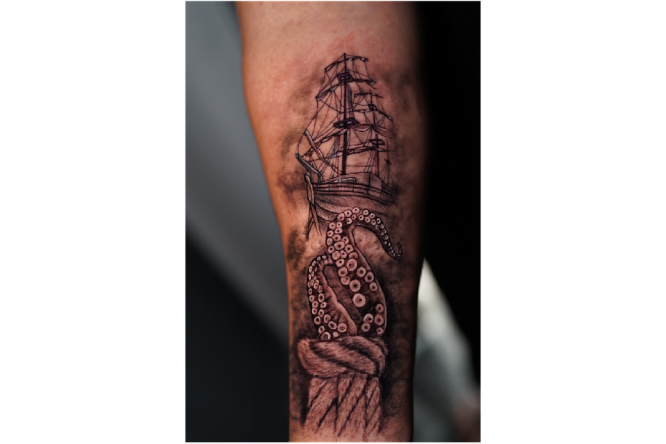 Image of a man's forearm with a maritime tattoo of a rope, octopus tentacles, a ship and a background in black, grey and white ink