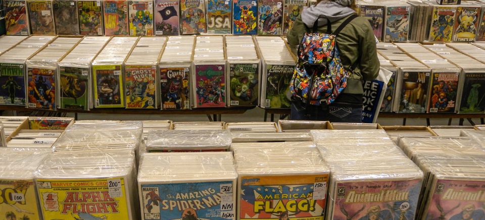 Attendee browsing through rows of bagged and boarded comic books displayed on tables at a convention, with colorful vintage and modern comic covers visible, including Spider-Man and American Flagg!.