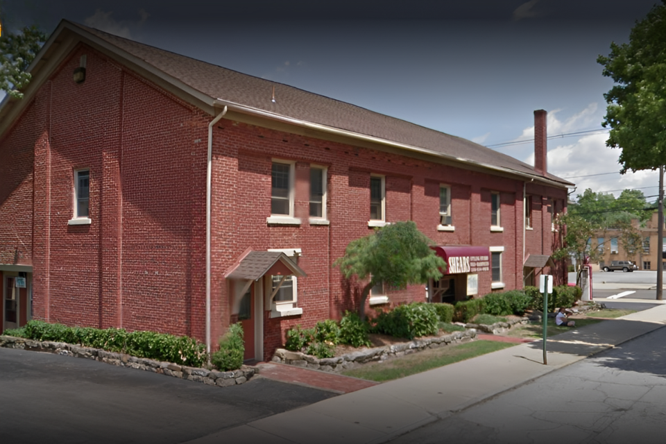 Exterior of the commercial building where Shears Wigs & Hairpieces Salon is located, featuring a brick façade, landscaped entrance, and sidewalk access.
