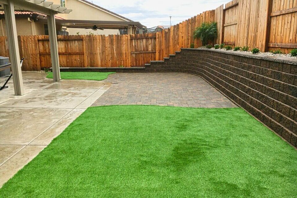 Modern low-maintenance backyard transformation featuring a tiered stone retaining wall, high-quality artificial turf, and a custom paver patio under a residential pergola.