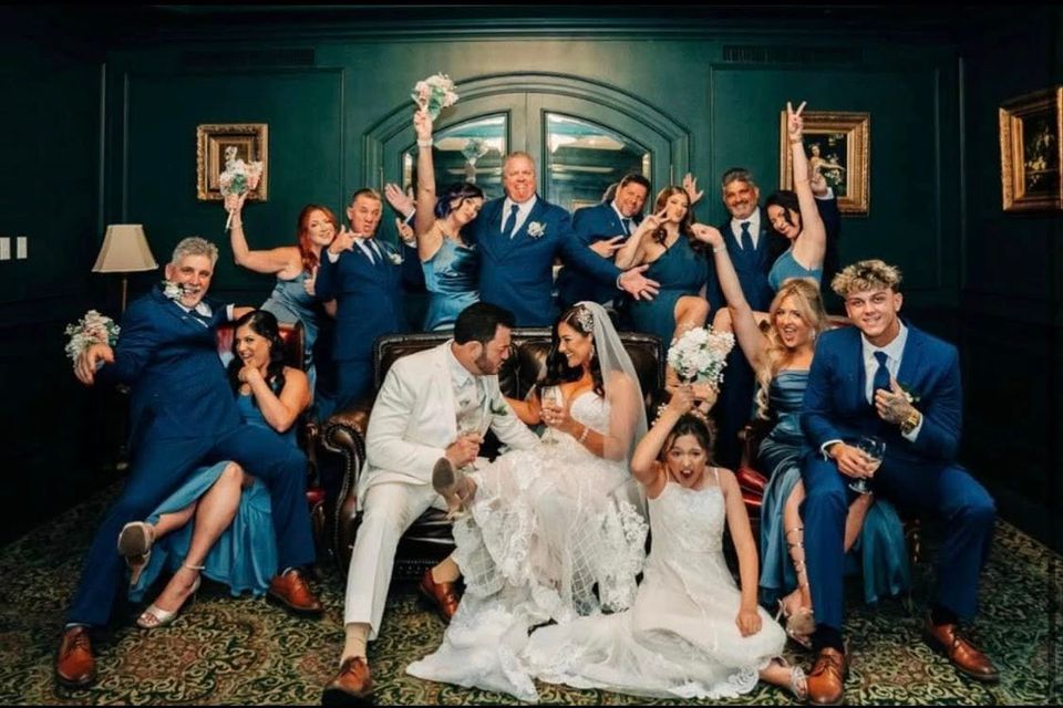 Bride and groom surrounded by a fun, energetic wedding party wearing coordinated blue tuxedos from New Orleans Tuxedo Inc., posing for a celebratory photo in an elegant indoor lounge.