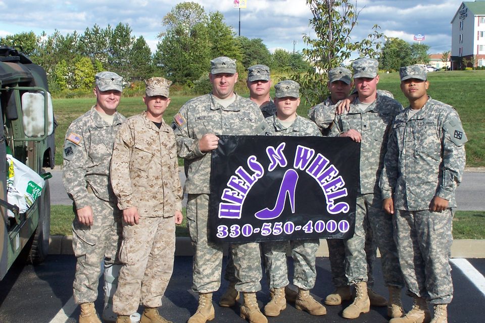 Group of U.S. service members holding a Heels 'N Wheels banner during a community support event.