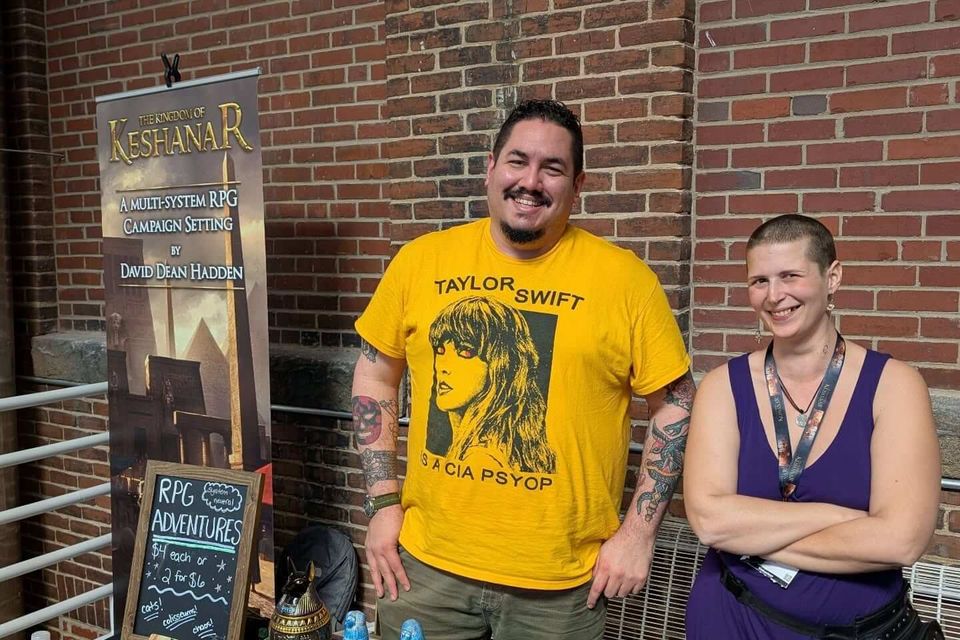 Two people smiling behind an RPG booth with Kingdom of Keshanar banner, Egyptian miniatures, and promotional materials