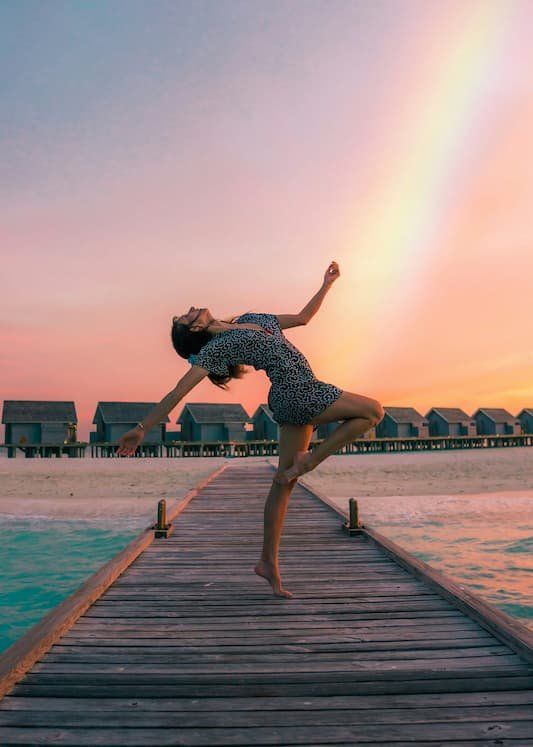 Person in a dancing position on a pier with a rainbow beaming down