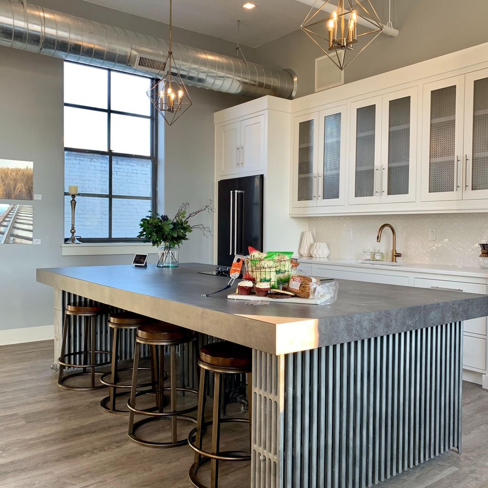 Modern industrial loft kitchen featuring an oversized matte grey concrete-look island with a fluted metal base and geometric gold pendant lighting—Artisan stone fabrication and custom surface installation by LuXe Design Tile and Granite inc.