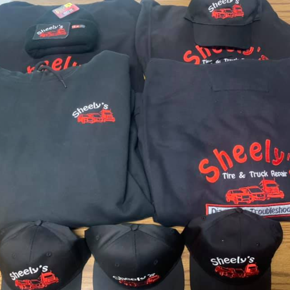 Custom embroidered Sheely's Tire & Truck Repair shirts, jackets, and hats displayed together on a table.
