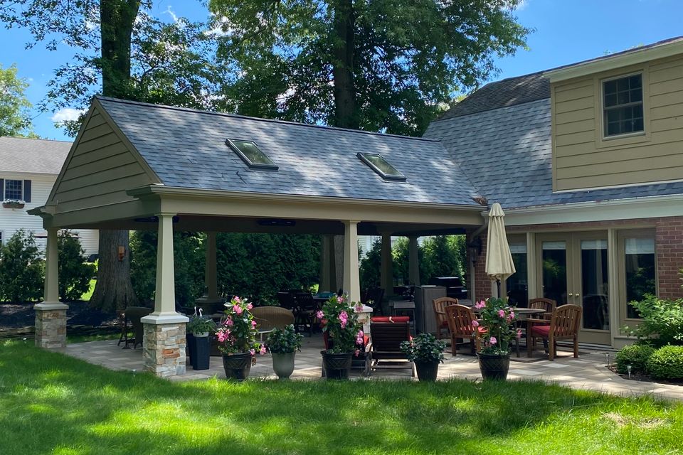 Covered backyard patio addition with stone columns, outdoor seating, dining area, and landscaped surroundings.