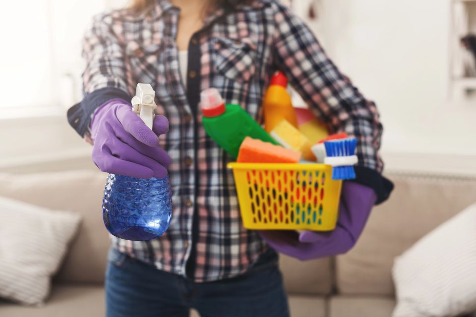 A woman holding a basket of cleaning supplies and is spraying a bottle of cleaner in the other hand with purple gloves on