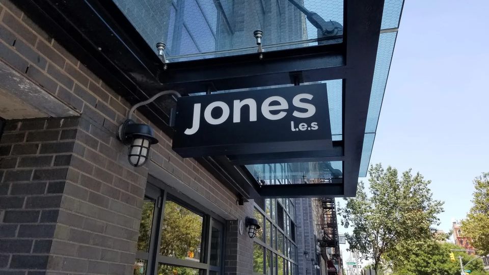 Black blade sign reading Jones L.E.S. hanging beneath a glass entrance canopy beside a wall light on the Lower East Side